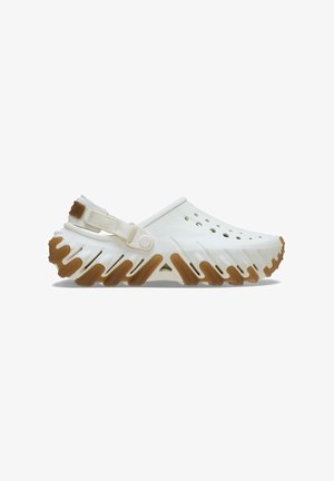 White plastic clogs with open-toe design, side ventilation holes, and a textured rubber sole featuring a zigzag pattern and brown accents.