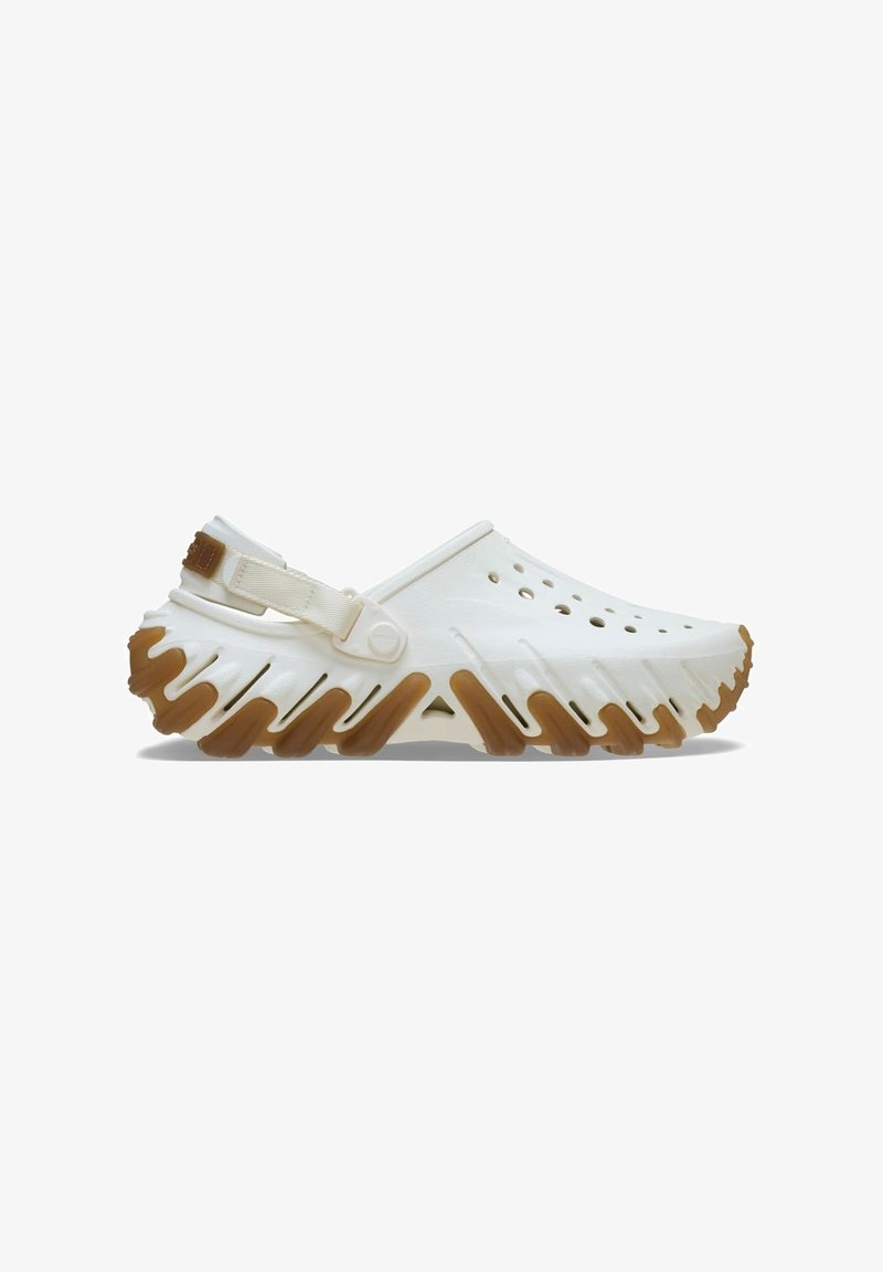 White plastic clogs with open-toe design, side ventilation holes, and a textured rubber sole featuring a zigzag pattern and brown accents.