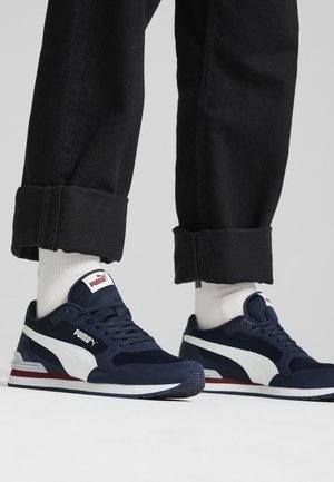 Osoba nosząca białe skarpetki i granatowe buty sportowe Puma z czarnymi podwiniętymi spodniami na szarym tle.