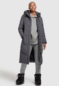 Langer grauer Puffermantel mit Kapuze, mit gestepptem Design, Seitenknöpfen und glatter Oberfläche. Über einem grauen Pullover und passenden Hosen getragen.