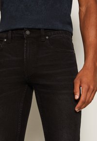 Man som bär åtsittande svarta jeans och en mörkblå skjorta, med en hand avslappnat vid sin sida mot en enkel bakgrund.