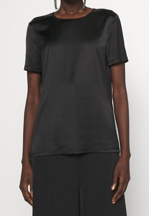 Personne portant un chemisier en satin noir uni à manches courtes avec un col rond et un pantalon noir, sur un fond blanc uni.