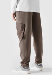 Cargo byxor i mjukt, brunt tyg med en avslappnad passform, med en stor sidoficka och elastiska muddar, i kombination med ljusgrå sneakers.
