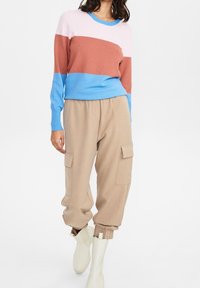 Bunter Pullover mit geripptem Muster in den Farben Pink, Braun und Blau. Hoch taillierte beige Cargo-Hose und weiße Stiefeletten.