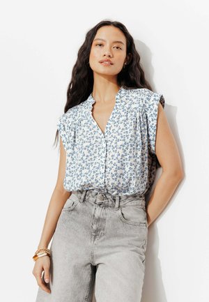 Femme aux longs cheveux foncés portant un chemisier blanc sans manches avec un imprimé floral bleu et un jean gris clair taille haute, debout contre un mur blanc.