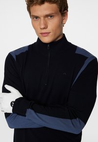 Svart tröja med hög krage, med blåa tyginslag på axlarna och ärmarna. Tillverkad av ett mjukt, texturerat stickat material.