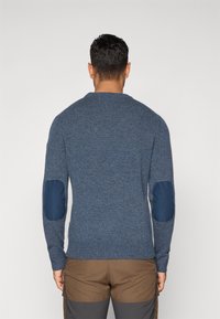 Bărbat văzut din spate, purtând un pulover din lână albastru cu patch-uri întunecate la coate și pantaloni maronii cu panouri gri.