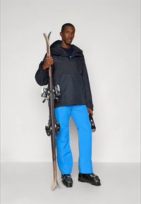 Άνδρας που φορά μαύρη τζακέτα και μπλε σκι παντελόνι κρατώντας σκι και γυαλιά σκι, στέκεται σε λευκό φόντο.