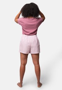 Lila Baumwollhemd mit kurzen Ärmeln, kombiniert mit hellrosa und weiß gestreiften Shorts. Shorts mit elastischem Bund. Einfarbiger Hintergrund.