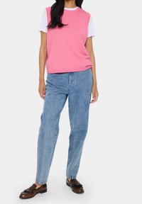 Kvinna som bär en rosa ärmlös jumper över en vit t-shirt, blå högmidjade jeans och bruna läder loafers stående mot en vit bakgrund.