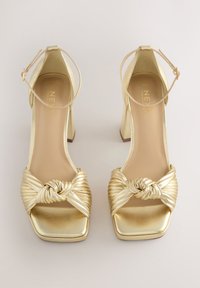 Zlaté sandále na podpätku so zaviazaným, plisovaným remienkom, štvorcovou špičkou a členkovým remienkom s kovovým leskom. Hladká textúra, široký podpätok.