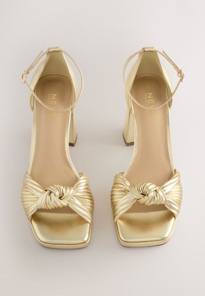 Złote sandały na obcasie z ozdobionym węzłem, plisowanym paskiem, kwadratowym noskiem i paskiem wokół kostki o metalicznym wykończeniu. Gładka tekstura, obcas typu klocek.