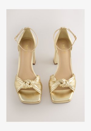 Złote sandały na obcasie z ozdobionym węzłem, plisowanym paskiem, kwadratowym noskiem i paskiem wokół kostki o metalicznym wykończeniu. Gładka tekstura, obcas typu klocek.