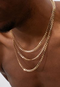 Goldene Kettenhalskette mit drei übereinander geschichteten Strängen, flachen Gliedern, glatter Textur und glänzender Oberfläche, die auf nackter Haut ruht.