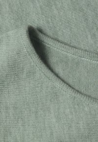 Tissu tricoté vert clair plié avec une texture côtelée visible et des coutures cousues formant un détail d'encolure ou de poignet.