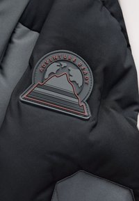 Tissu rembourré noir et gris avec un badge rond montrant des montagnes, un globe, et le texte "Adventure Ready" en lettres rouges.