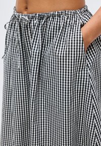 Γυναίκα που φοράει μαύρη και λευκή φούστα με σχέδιο gingham, με ελαστική μέση και κορδόνι, με το ένα χέρι στην πλαϊνή τσέπη.