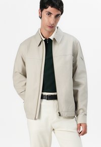 Beige ritsjack met kraag, met een soepele textuur en zijzakken. Gecombineerd met een donkergroene top en lichtgekleurde broeken.