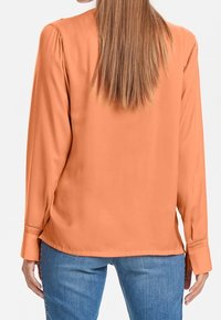 Blusa a maniche lunghe di colore corallo-arancio con una texture liscia, caratterizzata da cuciture della spalla plissettate e polsini con bottoni, abbinata a jeans blu.