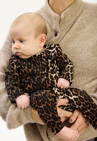 Leoparda raksta viengabala tērps brūnā, melnā un krēmkrāsā ar gariem piedurknēm un apaļu kaklu, ko tur cilvēks ar teksturētu bēšu džemperi.