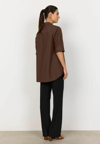 Chemise marron à boutons, à manches courtes, avec couture de yoke au dos et ourlet arrondi, assortie à un pantalon noir ample et des sandales.