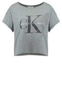 Šedé zkrácené tričko z bavlněné směsi, které má velké černé logo "CK" a text "Calvin Klein Jeans". Krátké volné rukávy, kulatý výstřih.