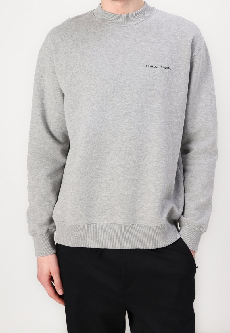 Homme en sweat-shirt gris clair à col rond avec un petit logo noir "SAMSOE SAMSOE" sur la poitrine, portant un pantalon noir, debout devant un fond uni clair.
