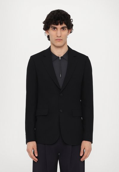 Blazer noir avec deux poches avant, revers crantés et fermeture à deux boutons, porté sur un haut zippé gris foncé. Texture lisse.