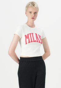 Hvit, kort t-skjorte med "MILAN" i fet rød skrift. Korte ermer, tettsittende design, kombinert med svarte bukser med elastisk midje.