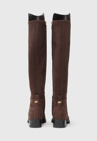 Knee-high boots made of brown suede dengan desain ramping, menampilkan aksen logo emas yang halus dan interior kulit hitam. Penutupan ritsleting di sisi.