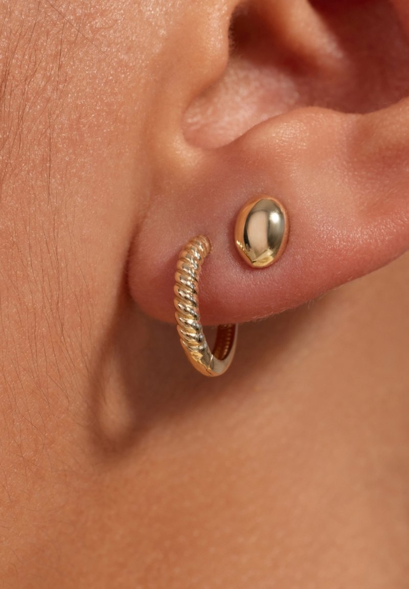 Goldfarbene Ohrringe mit einem gewundenen Ringdesign und einem glatten, ovalen Stecker mit reflektierender Oberfläche, vor einem Hautton.