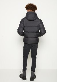 Sort pufferjakke med hætte, kviltet design og elastiske manchetter, båret med sorte jeans og ankelstøvler. Stoffet ser glat og fyldigt ud.