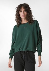 Zelená mikina s volným střihem, ribovaný lem a dlouhými rukávy. Na hrudi se nachází logo "Goddess" a černé detaily podél rukávů.