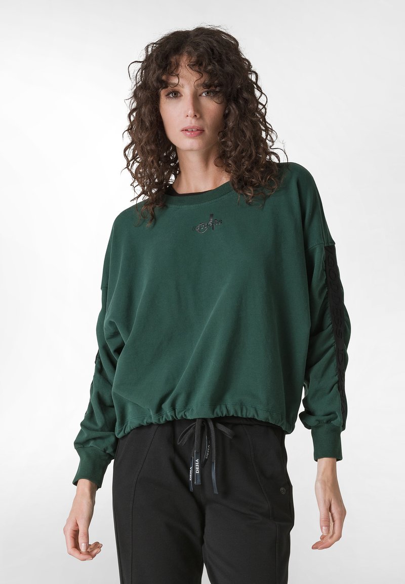 Zelená mikina s volným střihem, ribovaný lem a dlouhými rukávy. Na hrudi se nachází logo "Goddess" a černé detaily podél rukávů.