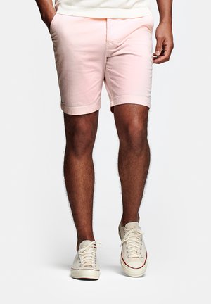 Man die lichtroze korte broek en witte sneakers draagt, staand met één hand in de zak tegen een effen witte achtergrond.