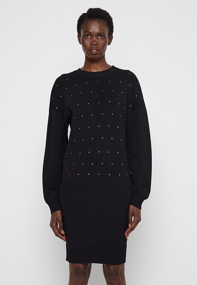 Frau, die ein schwarzes, langärmliges Strickkleid mit kleinen metallischen Nieten im oberen Bereich trägt, steht vor einem schlichten hellen Hintergrund.