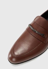 Brown leather loafer with a pointed toe, featuring a metal chain accent across the front and stitched detailing along the seams.