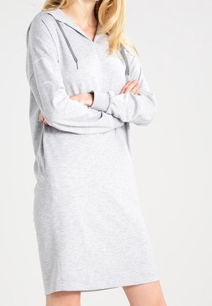 Kobieta w jasnoszarej sukience z kapturem z długim rękawem i sznurkami, ze skrzyżowanymi ramionami, na tle jednolitego, jasnego tła.