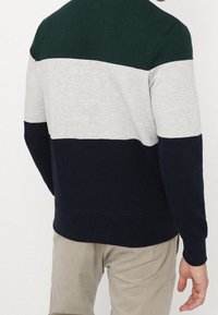 Hombre vistiendo un suéter de bloques de color con franjas horizontales en verde oscuro, gris claro y azul marino, acompañado de pantalones beige.