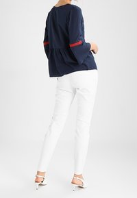 Top strutturato blu navy con rifiniture rosse sulle maniche, abbinato a pantaloni bianchi slim-fit e sandali con tacco alto, visto da dietro.