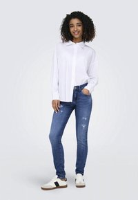 Camisa blanca de manga larga, con botones, combinada con vaqueros rotos de color azul oscuro y zapatillas blancas con detalles en negro. Outfit casual.