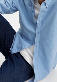 Camicia azzurra chiara in cotone, con una texture leggermente stropicciata. Abbinata a jeans scuri in denim e una maglietta bianca sotto.