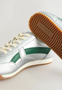 Baskets de sport argentées avec des accents verts, semelle en caoutchouc texturé, lacets crème et un design épuré. Détail de marque notable sur la semelle.