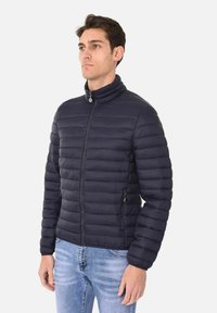 Giacca a vento blu navy con colletto alto, design a cannelloni orizzontali, tasche con zip sul davanti e polsini elasticizzati, indossata sopra jeans azzurri chiari.