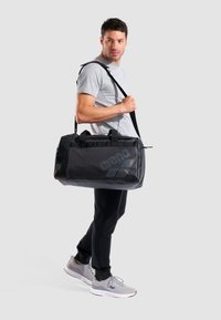 Bolsa de deporte negra con un acabado texturizado, que cuenta con un logo prominente, asas dobles y una correa de hombro desmontable.