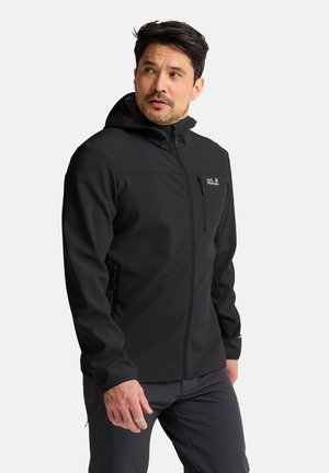 Man draagt een zwarte softshell jas met capuchon, rits en borstzak, kijkt naar links.