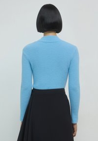 Blauer strukturierter Pullover mit hohem Kragen und langen Ärmeln, kombiniert mit einem schwarzen Rock. Der glatte Stoffkontrast hebt die Designelemente hervor.