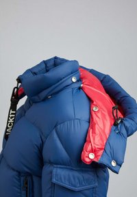 Blauwe pufferjack met een hoge kraag, voorzien van een contrasterende rode accenten op één schouder, drukknoopsluitingen en details van voorzakken.