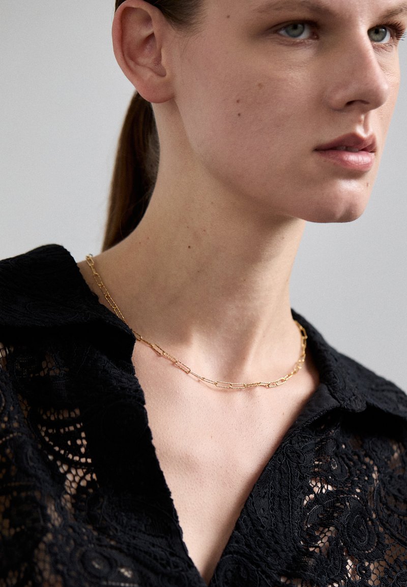 Kuldne kettkaelake, millel on piklikud, ristkülikukujulised lingid. Kantud musta pitsilise kaelusega särgi peal, näidates siledat ja poleeritud viimistlust.