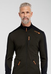 Lynlukkede jakke i mørkegrøn med sorte ærmer. Har orange lynlåsdragter og et logo på venstre skulder. Fremstillet af et strækstof.